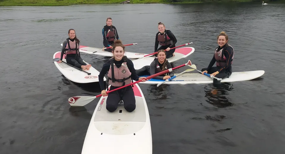 Studenti del Breifne College con la muta indossata stanno sui paddleboard in acqua circondati dal verde della natura.