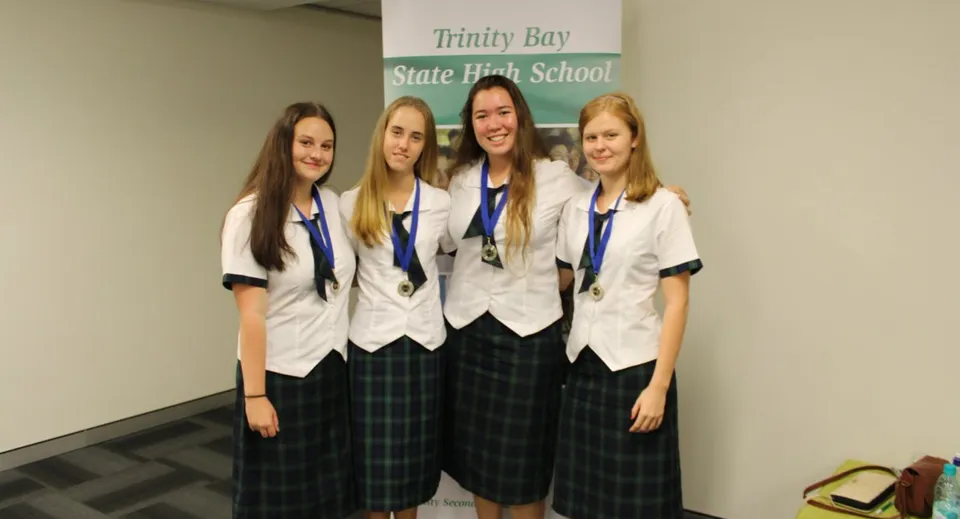 Quattro studentesse in uniforme scolastica sono insieme davanti al cartello della Trinity Bay State High School.