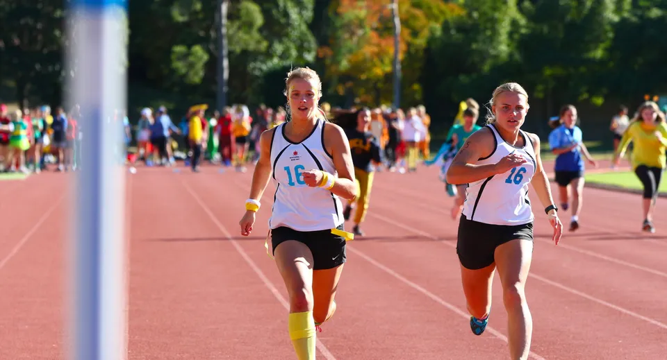 Due atlete corrono sulla pista di atletica nel campus della Kincoppal-Rose Bay davanti a spettatori e alberi.