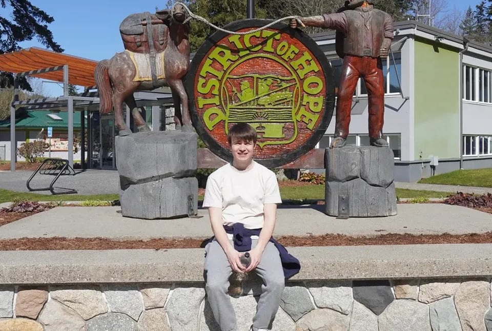 Stefan sitzt vor dem Schild „District of Hope“ mit Holzskulpturen in Kanada