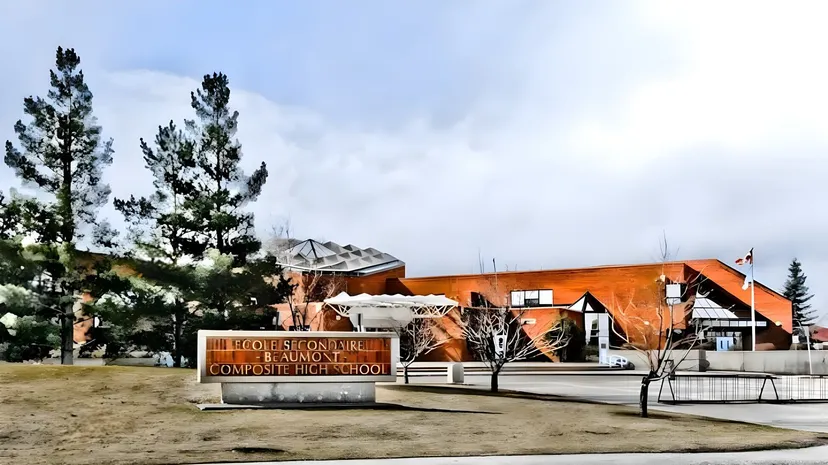 L'edificio moderno di colore arancione della École Secondaire Beaumont Composite High School sorge circondato da pini e da un prato sotto un cielo nuvoloso.