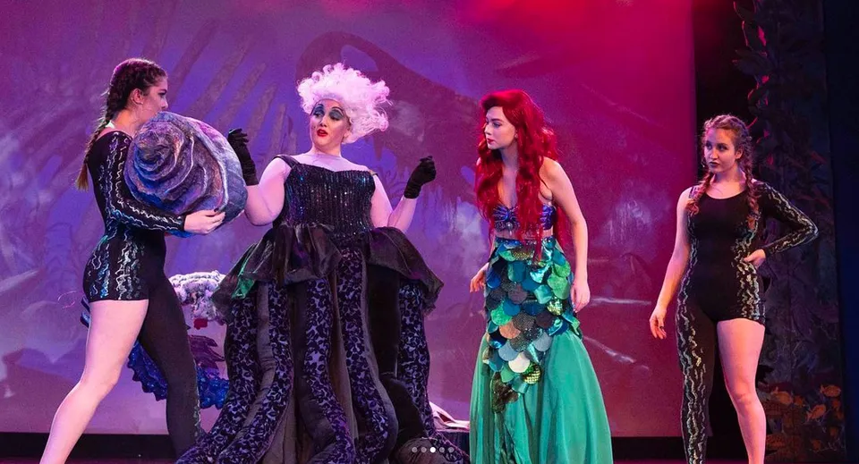 Quattro studenti della Claremont Secondary School indossano costumi colorati su un palco con una vivace scenografia teatrale.