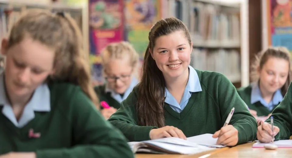 Una studentessa sorridente della Rathdown School indossa la divisa scolastica verde e scrive a un banco.