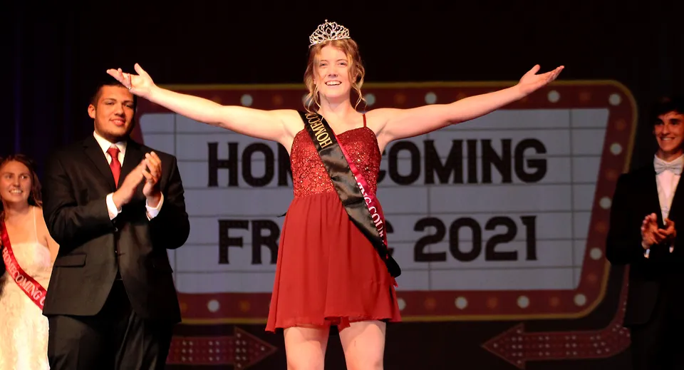 Una giovane donna in abito rosso e corona sta a braccia alzate durante l'evento di Homecoming alla Fox Valley Lutheran High School.