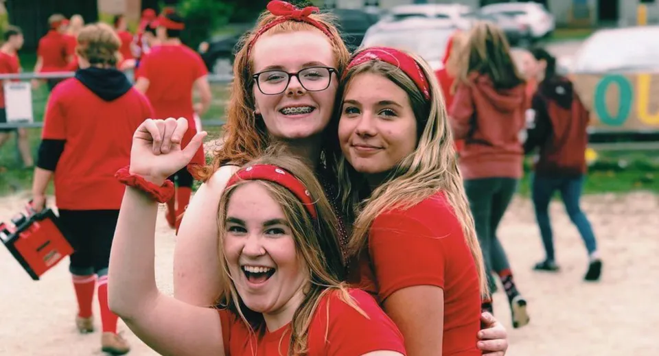 Tre studentesse del Stayner Collegiate Institute indossano magliette rosse e sorridono davanti a una folla di persone.