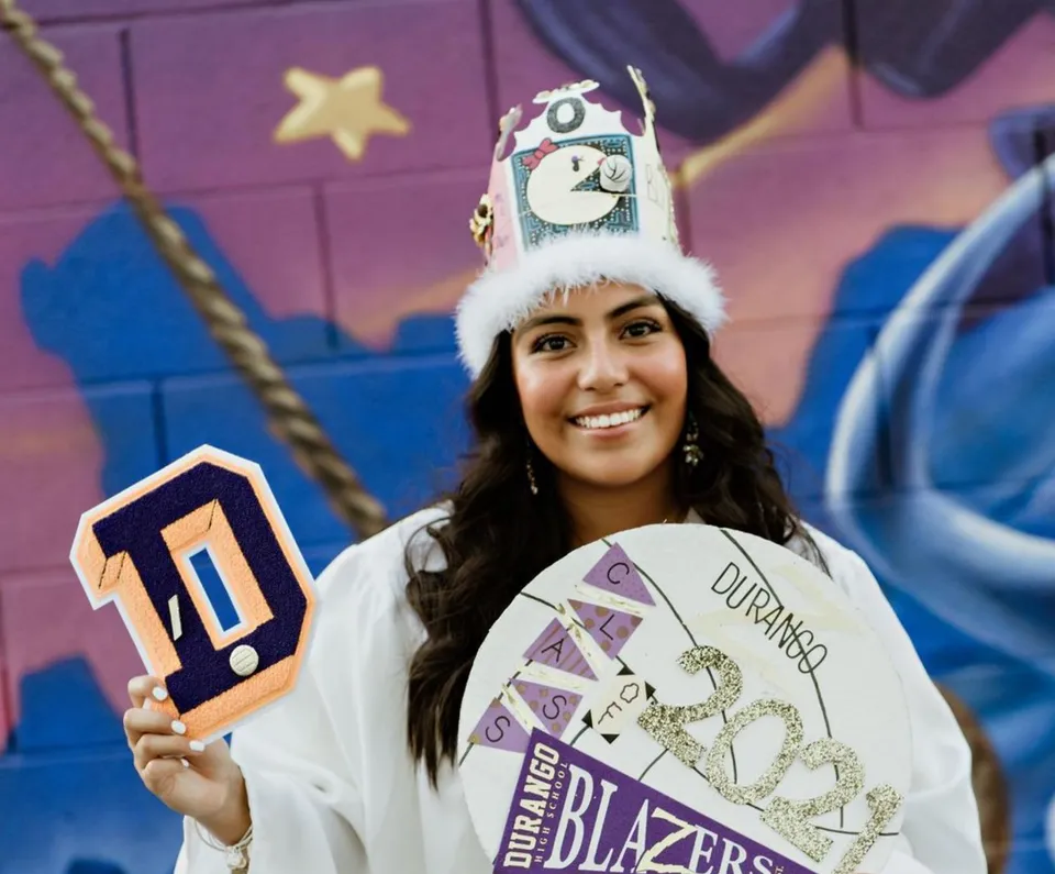 Una studentessa della Durango High School con un cartello con il numero 1 davanti a un muro dipinto con graffiti colorati.