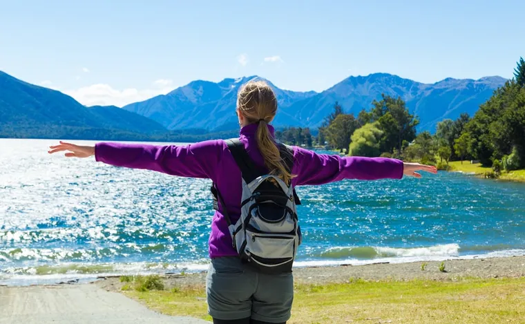 Frau mit Rucksack breitet am Ufer eines Bergsees die Arme aus