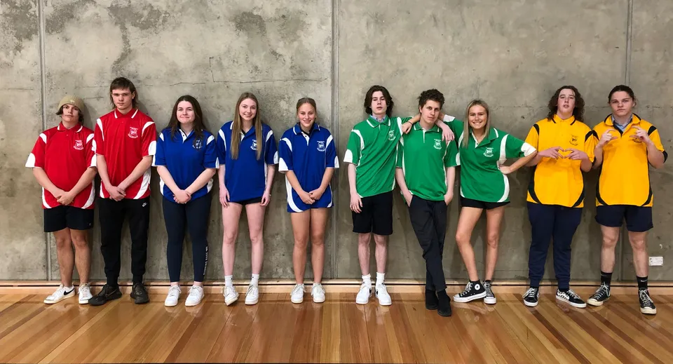 Un gruppo di giovani atlete si trova in diverse divise sportive colorate davanti a una parete di cemento nel terreno della Roxby Downs Area School.