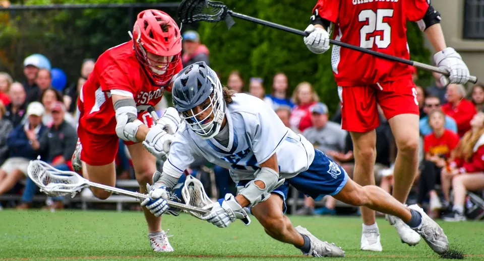 Due giocatrici in rosso e bianco giocano a lacrosse sul campo della Wyoming Seminary College Preparatory con spettatori sullo sfondo.