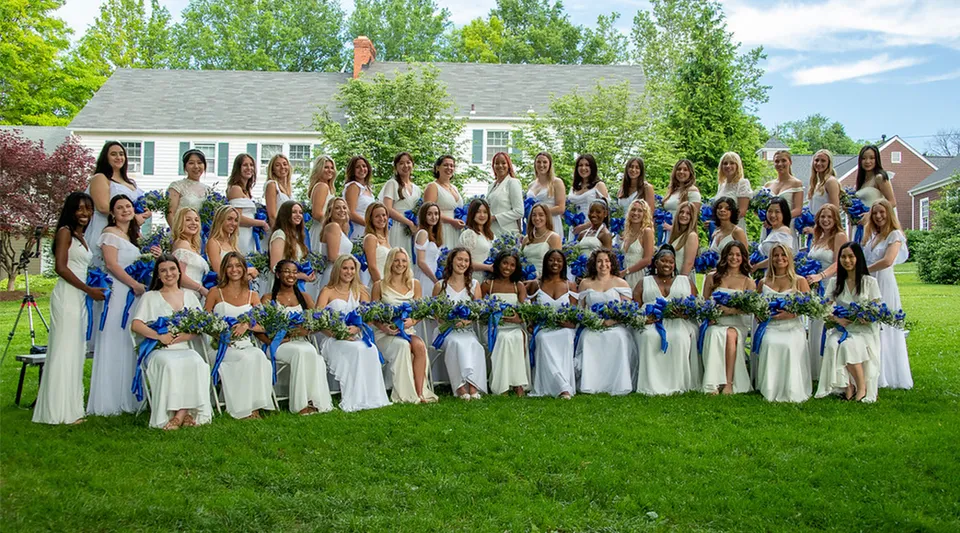 Un grande gruppo di donne in abito bianco con dettagli blu è in piedi su un prato davanti a una casa nel terreno della Garrison Forest School.