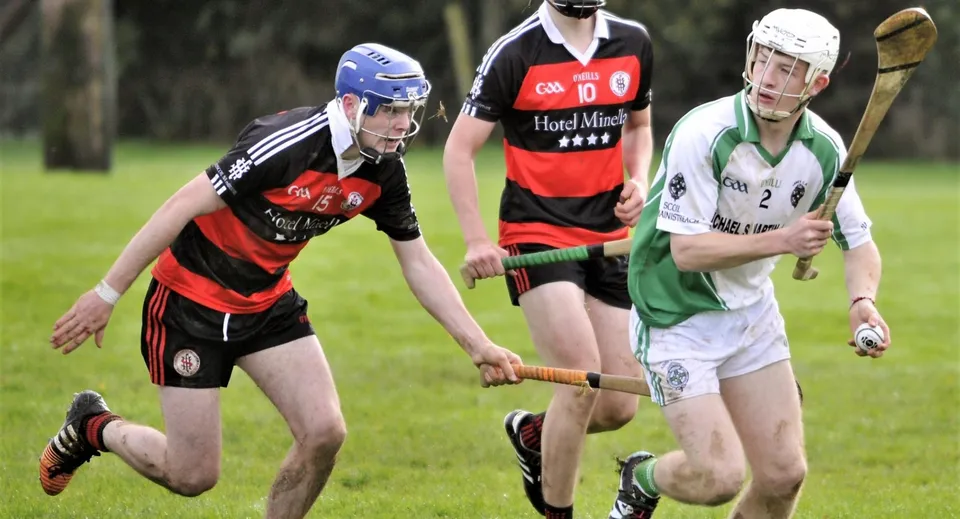 Tre studenti della Scoil Aireagail giocano a hurling con caschi e bastoni su un campo in erba.
