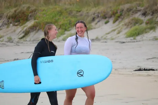 Zwei Austauschschülerinnen tragen ein Surfbrett am Strand