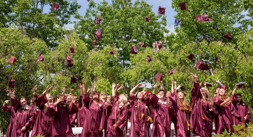 Un gruppo di diplomati della Kents Hill School in toga color bordeaux festeggia il diploma sotto alberi verdi.