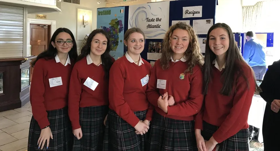 Cinque studentesse della Sacred Heart Secondary School Clonakilty sono insieme nel corridoio della scuola davanti a cartelloni colorati.
