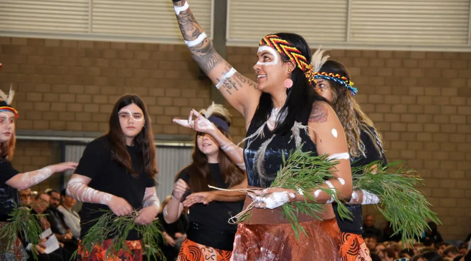 Un gruppo di studenti del Gilmore College si esibisce in costumi variopinti di ispirazione tribale su un palco davanti a una parete di mattoni.