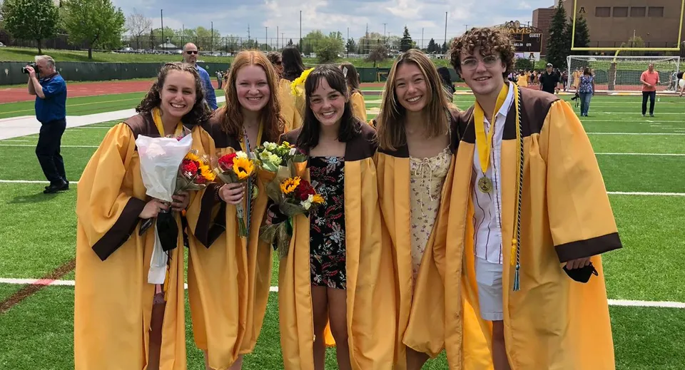 Un gruppo di giovani donne in toga da diploma tiene dei fiori sul campo sportivo delle Rochester Community Schools.