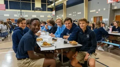 Estudiantes de Liberty Christian Academy están sentados juntos en la cafetería y comen una comida bajo canastas de baloncesto.