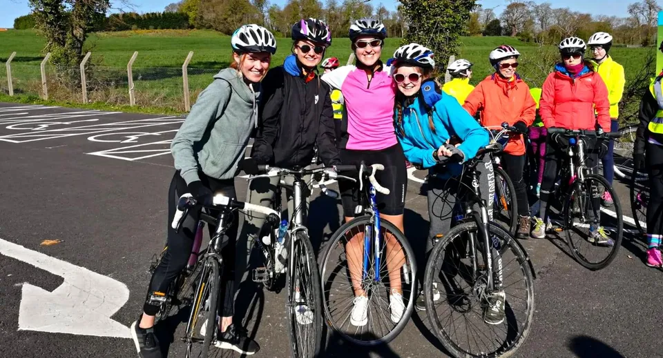Un gruppo di studenti della St. Raphaela’s Secondary School con abbigliamento da ciclismo colorato posa su una strada davanti a un prato verde.