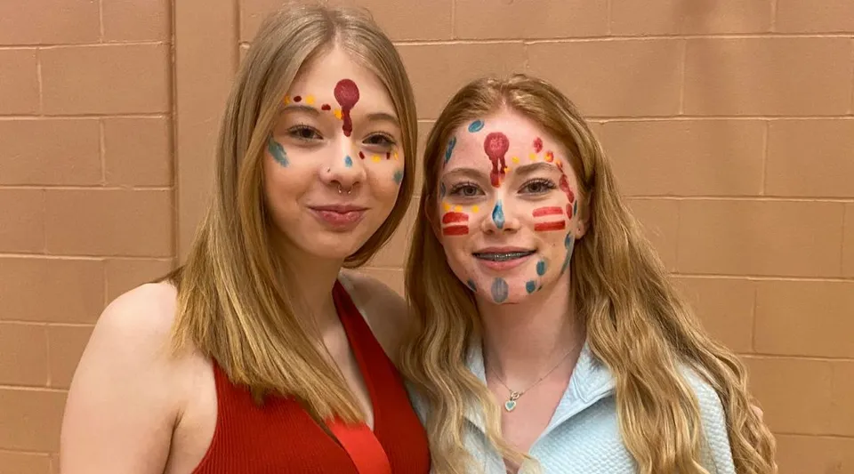 Due studentesse della Mobile Central High con il viso dipinto a colori sorridono davanti a una parete beige.