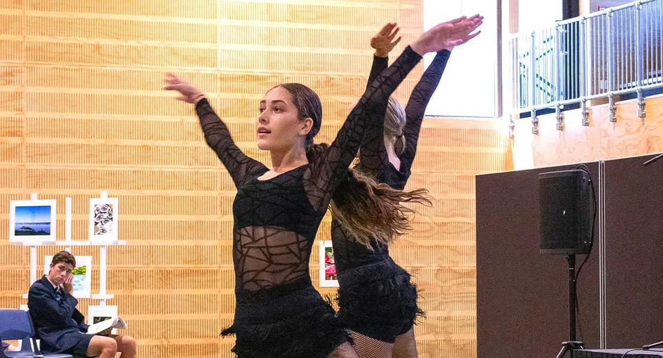 Una donna con un abito nero con piume si esibisce sul palco dell'ACG Tauranga, sullo sfondo si vedono una parete in legno e opere d'arte incorniciate.