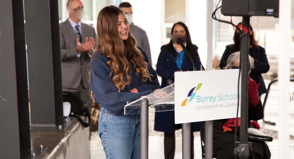 Un gruppo di persone si trova davanti a un cartello con la scritta Surrey Service sul terreno della Earl Marriott Secondary School.