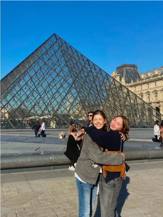 Zwei Mädchen umarmen sich lächelnd vor der Louvre-Pyramide in Paris