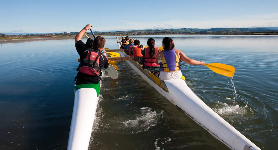 Studenti del William Colenso College che remano insieme su una canoa colorata su un lago tranquillo con montagne sullo sfondo.