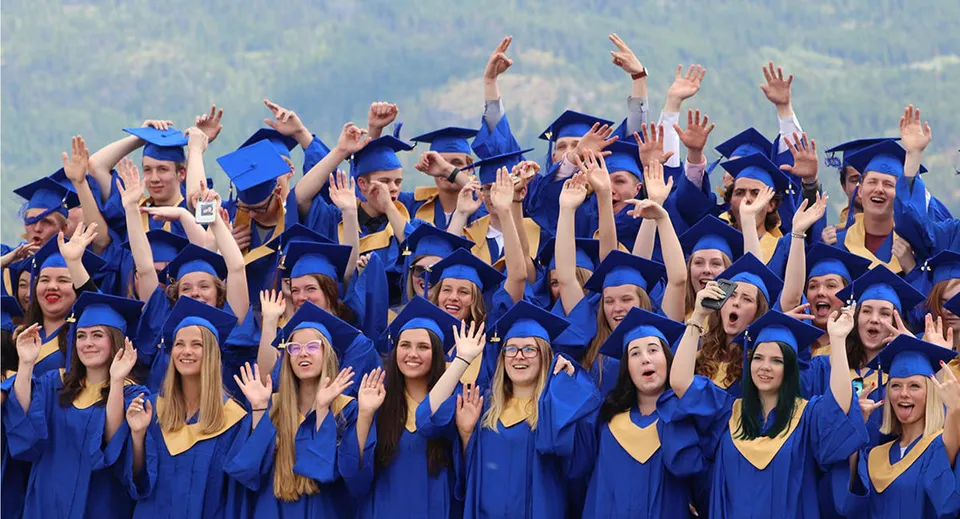 I diplomati della Kalamalka Secondary School festeggiano il loro diploma in toga blu e gialla davanti a un paesaggio montuoso.