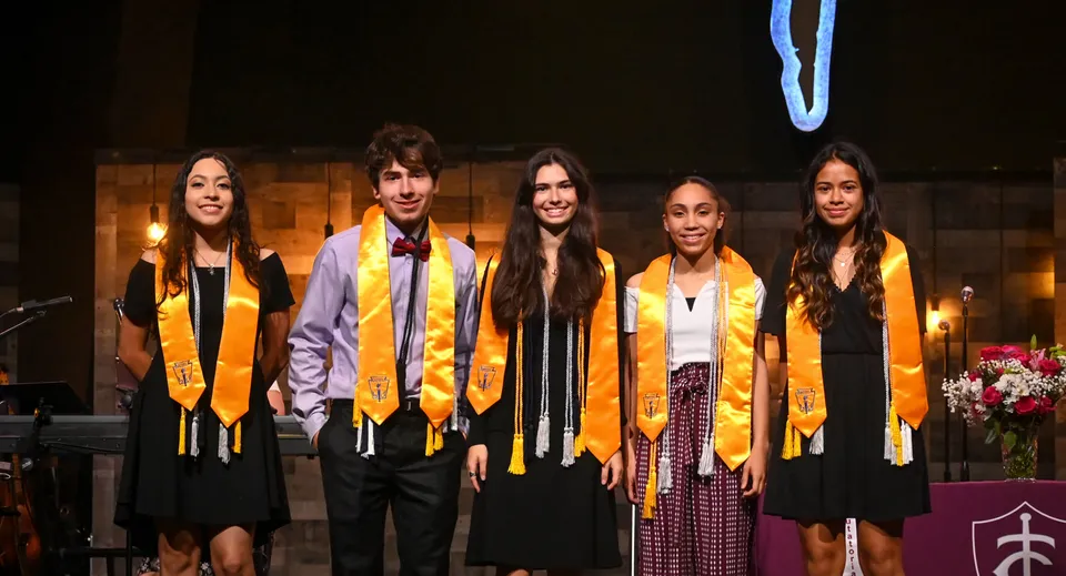 Un gruppo di cinque studenti e studentesse in abito da diploma sta in piedi con candele e fiori sullo sfondo nel campus della Trinity Christian Academy.