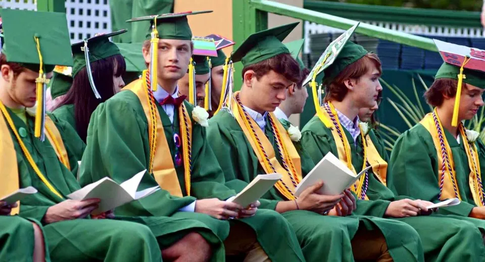 Un gruppo di diplomati in toga verde mostra i propri diplomi davanti a uno sfondo colorato nel campus della St. Johnsbury Academy.