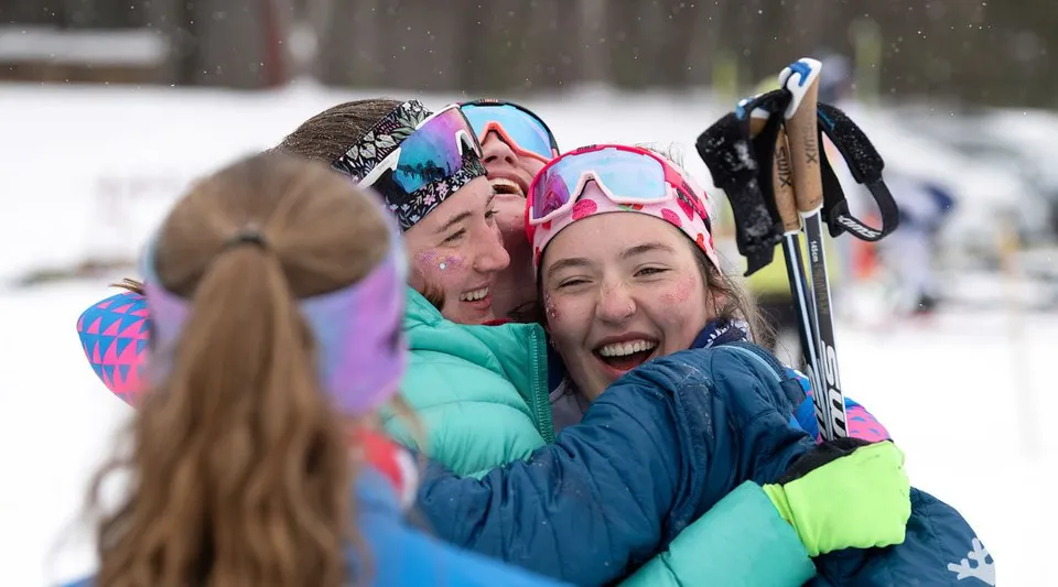 Studentesse della Gould Academy con occhiali da sci e abbigliamento invernale si abbracciano sorridenti sulla neve.