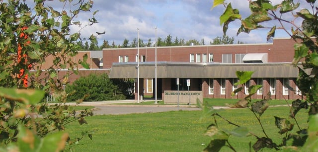 El edificio de ladrillo con entrada cubierta en el terreno de la Bernice McNaughton High School está rodeado de árboles y césped.