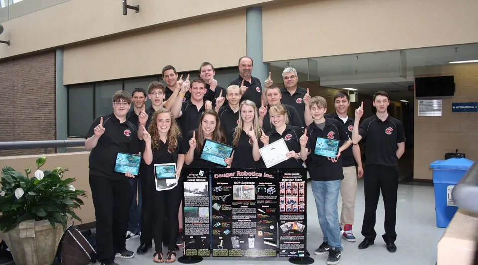 Un gruppo di studenti si trova davanti all'edificio della Clarenville High School e tiene in mano vari oggetti e un cartello.