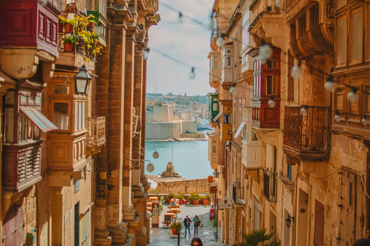 Calle estrecha de La Valeta con balcones de madera y el puerto al final de la bajada