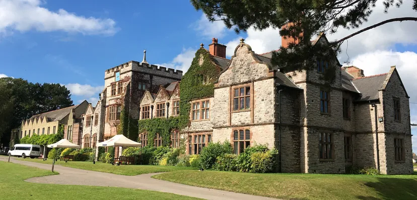 L'edificio storico con facciata in pietra della Ruthin School è circondato da erba verde e alberi sotto un cielo blu.