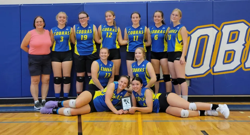 Una squadra femminile di pallavolo posa insieme sul campo da basket della Willow Creek Composite High School davanti a un grande logo OB.