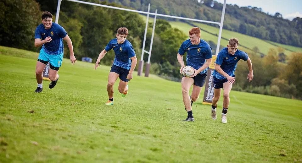 Studenti delle Haberdashers' Monmouth Schools corrono su un campo da gioco tra verdi colline e alberi.