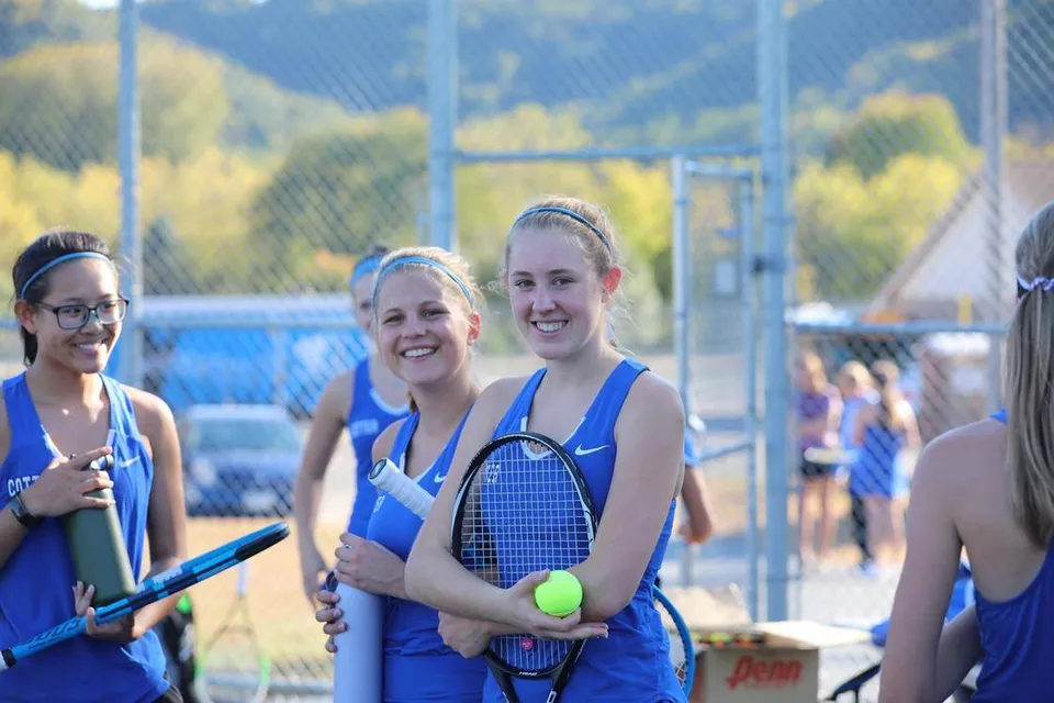 Tre studentesse in abbigliamento sportivo con racchette da tennis davanti al campo da tennis della Cotter Schools con montagne sullo sfondo.