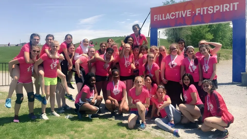 Un grupo de alumnas de la Riverside Secondary School posa con uniformes idénticos de color rosa y rojo frente a las letras Fillactive Fitspirit en un campo.