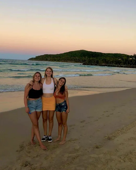 Tres chicas abrazadas en la playa al atardecer, con el mar y una colina verde al fondo