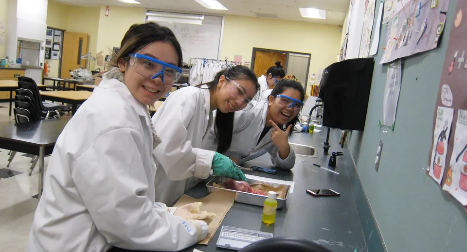 Tre studenti della A.R. MacNeill Secondary School lavorano insieme con camici bianchi da laboratorio presso una postazione di lavoro.