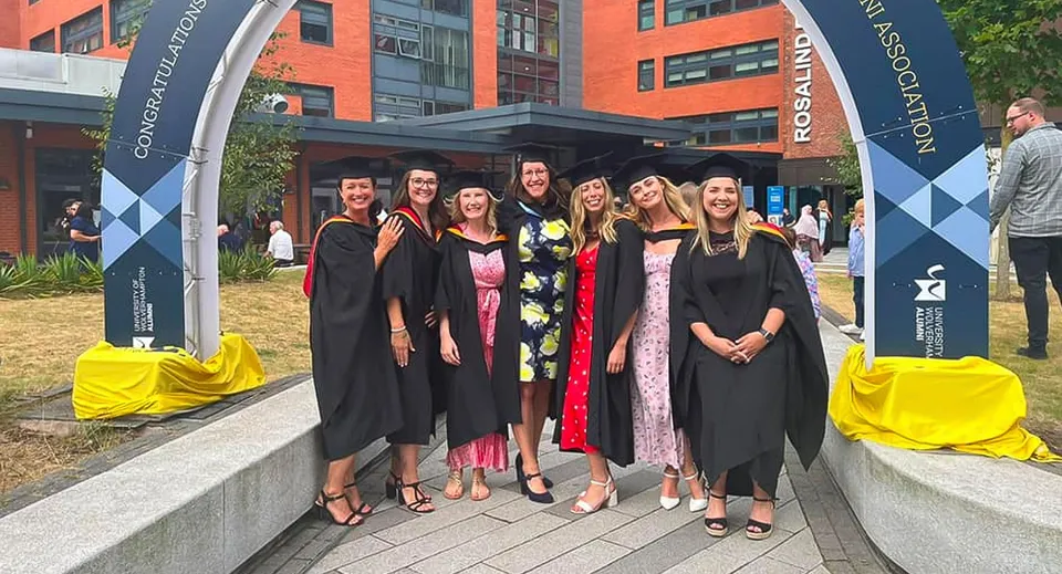 Gli studenti del Bournemouth & Poole College si trovano in toga da diploma davanti a un edificio universitario con striscioni sullo sfondo.