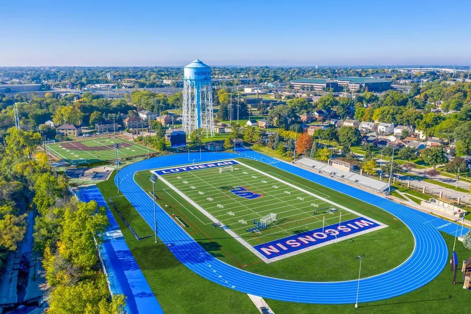 Il grande campo da football con la pista blu nel campus della Wisconsin Lutheran High School mostra sullo sfondo lo skyline della città con un'alta torre idrica.