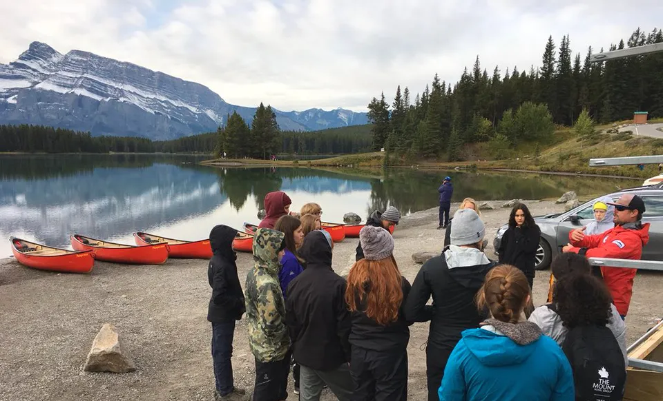 Un gruppo di studenti sulla riva di un lago con montagne e bosco sullo sfondo nel terreno della Canmore Collegiate High School.