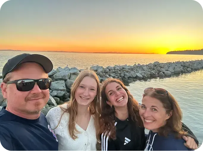 Selfie di famiglia sorridente al tramonto davanti al mare, con una scogliera di pietre sullo sfondo