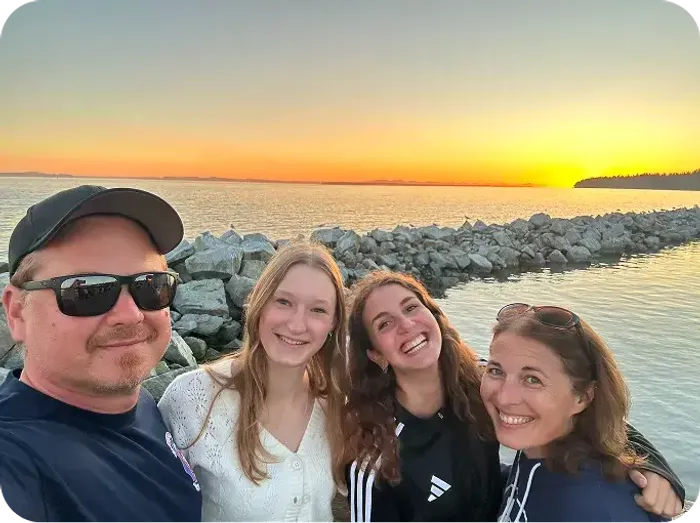 Selfie di famiglia sorridente al tramonto davanti al mare, con una scogliera di pietre sullo sfondo