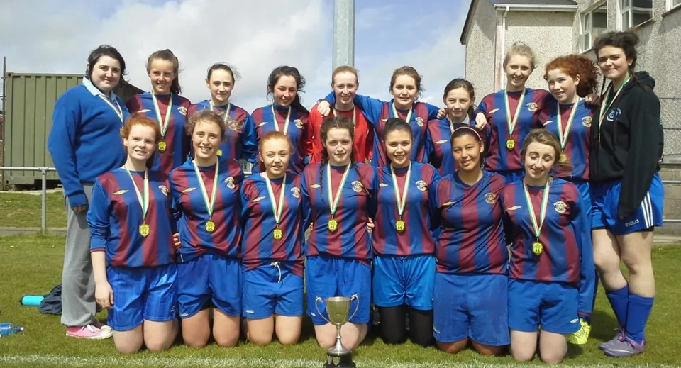 Un gruppo di studentesse della Eureka Secondary School posa insieme in maglie blu e rosse su un campo da calcio.