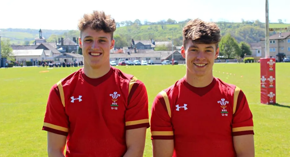 Due studenti del Llandovery College con la maglia sportiva rossa e l'emblema della bandiera del Wales si trovano su un campo erboso con vista su una città sullo sfondo.
