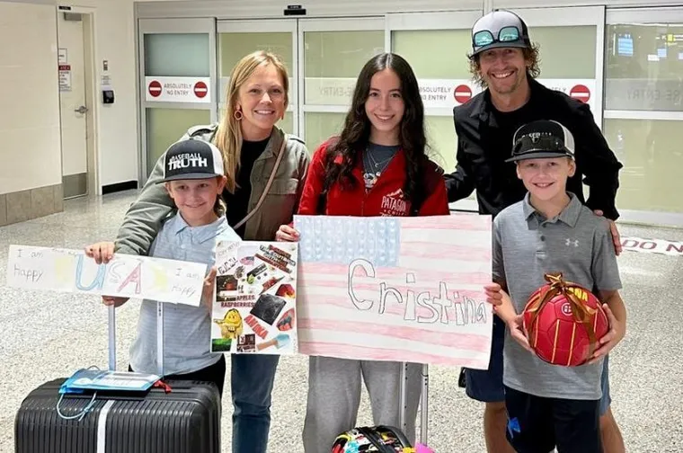 Familia sonriente posa en un aeropuerto sosteniendo carteles de bienvenida para una joven