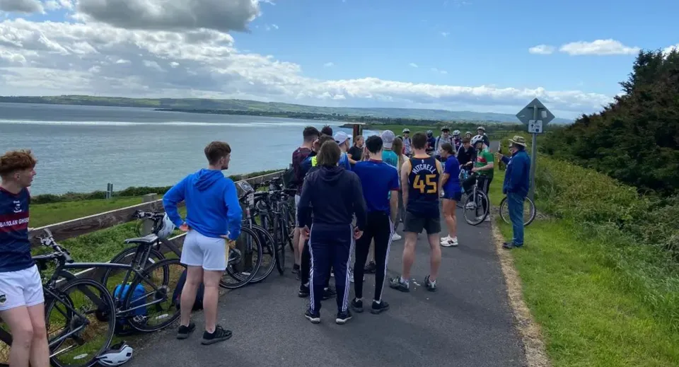 Un gruppo di studentesse e studenti della Ballingarry Presentation School si trova su un sentiero con vista su un lago e colline boscose.
